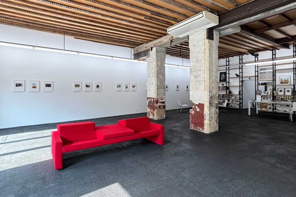La grande salle de la galerie : plafond de poutres, piliers de pierre, un canapé rouge face à un mur de photographies encadrées, la bibliothèque au fond.